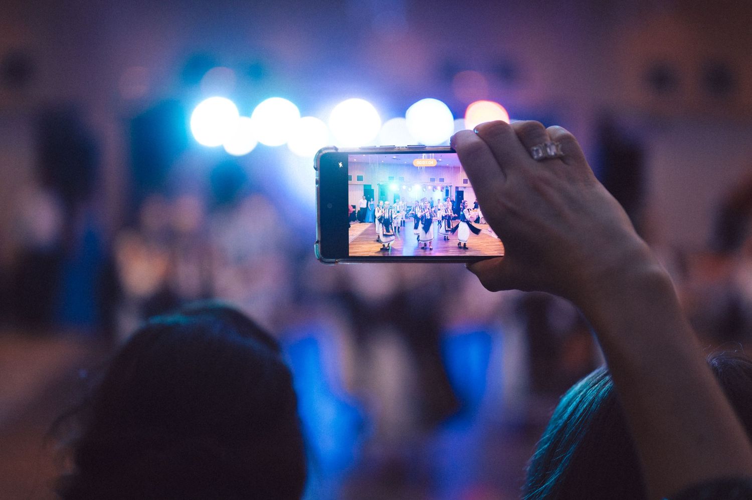 Someone holds up a smartphone to capture video of a concert or event with blue stage lights in the background.