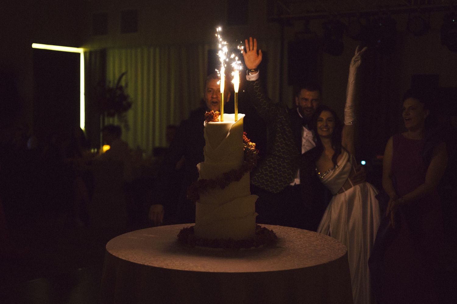 A wedding cake with sparkler fountains glows in a darkened reception room.