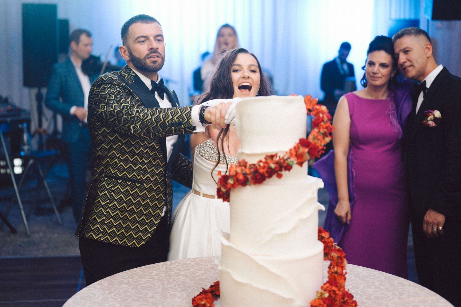 Couple cuts their elegant white wedding cake decorated with red floral accents.