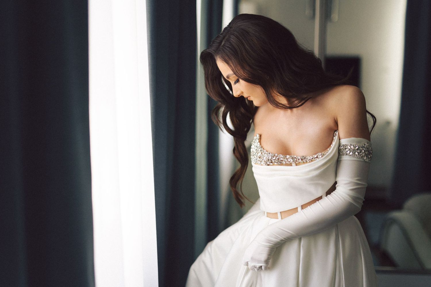An elegant off-shoulder wedding dress with delicate embellishments is showcased in a romantic window portrait.