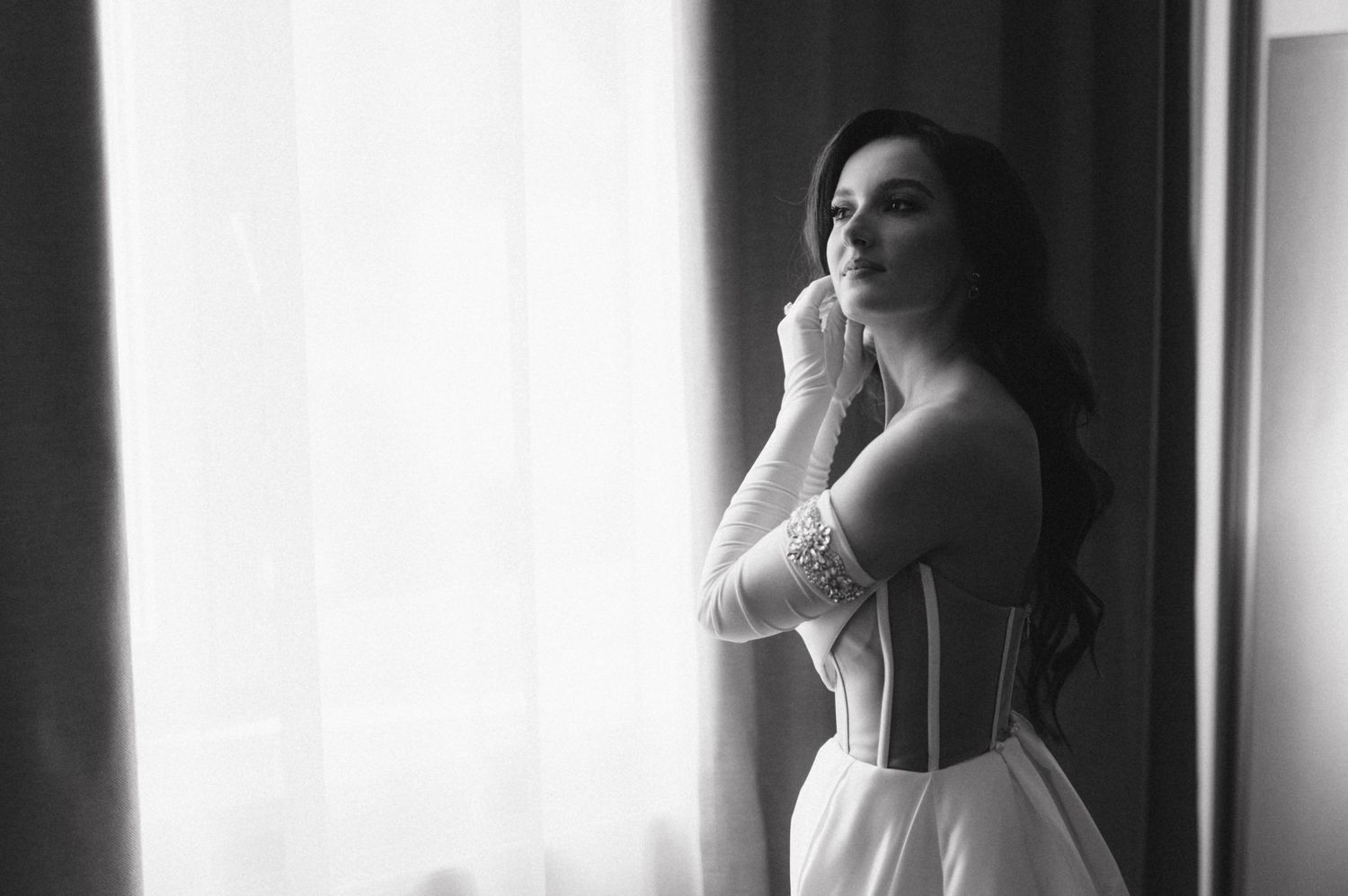 A black and white bridal portrait shows an elegant figure in a corseted dress near a bright window.