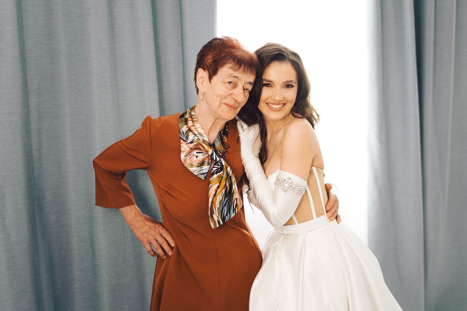 A person in a red dress poses with someone wearing a white wedding gown against a grey backdrop.