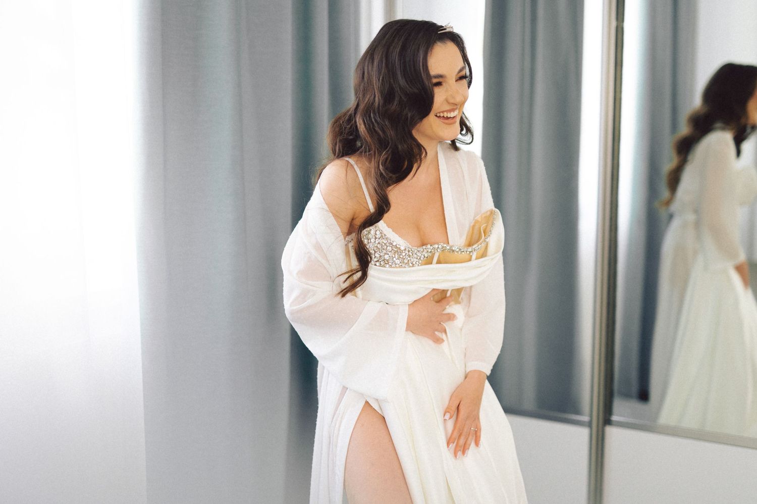 Woman in white robe reflected in mirror during bridal preparation.