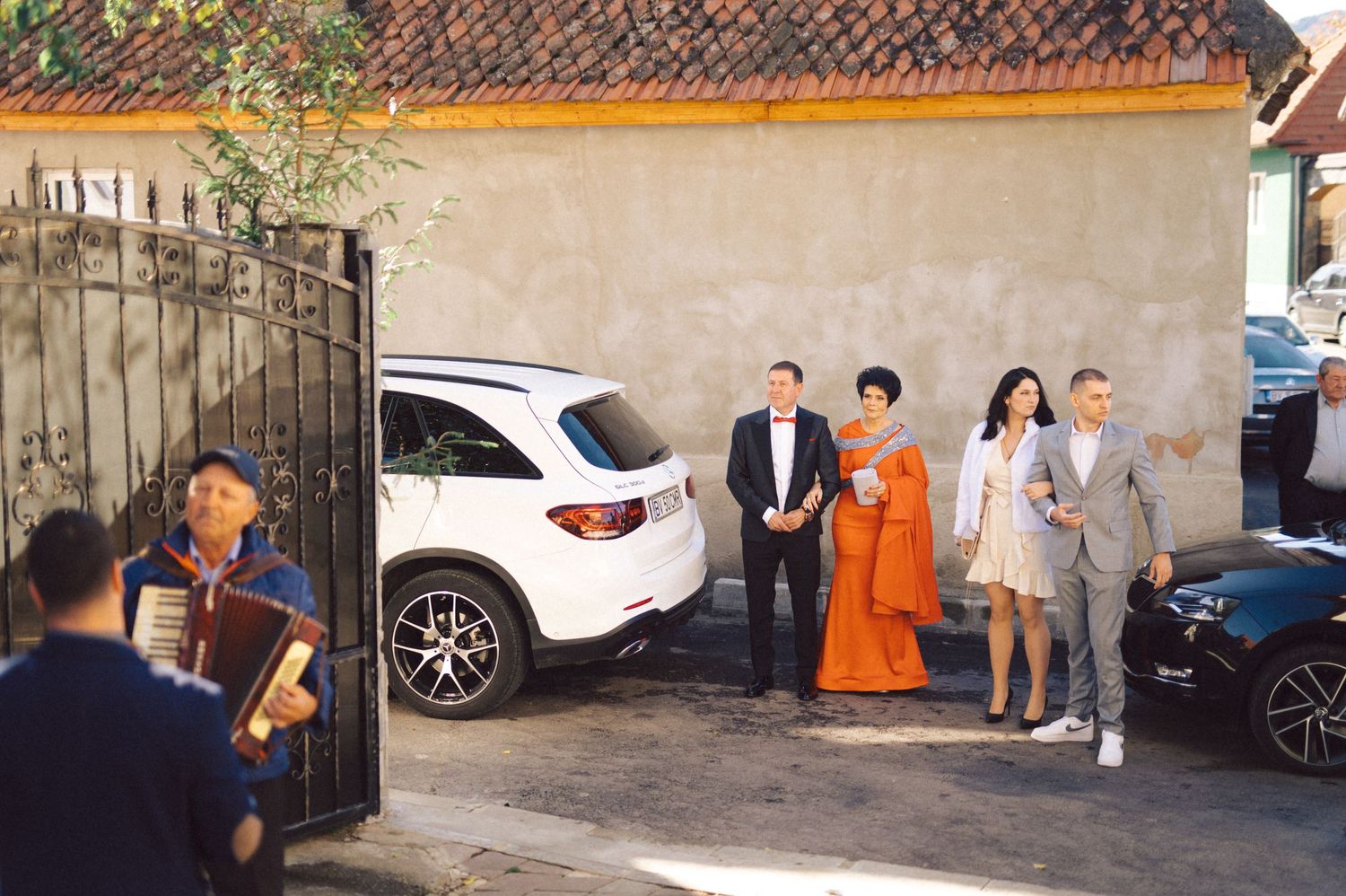 A wedding party arrives at a venue entrance with parked cars and iron gates.