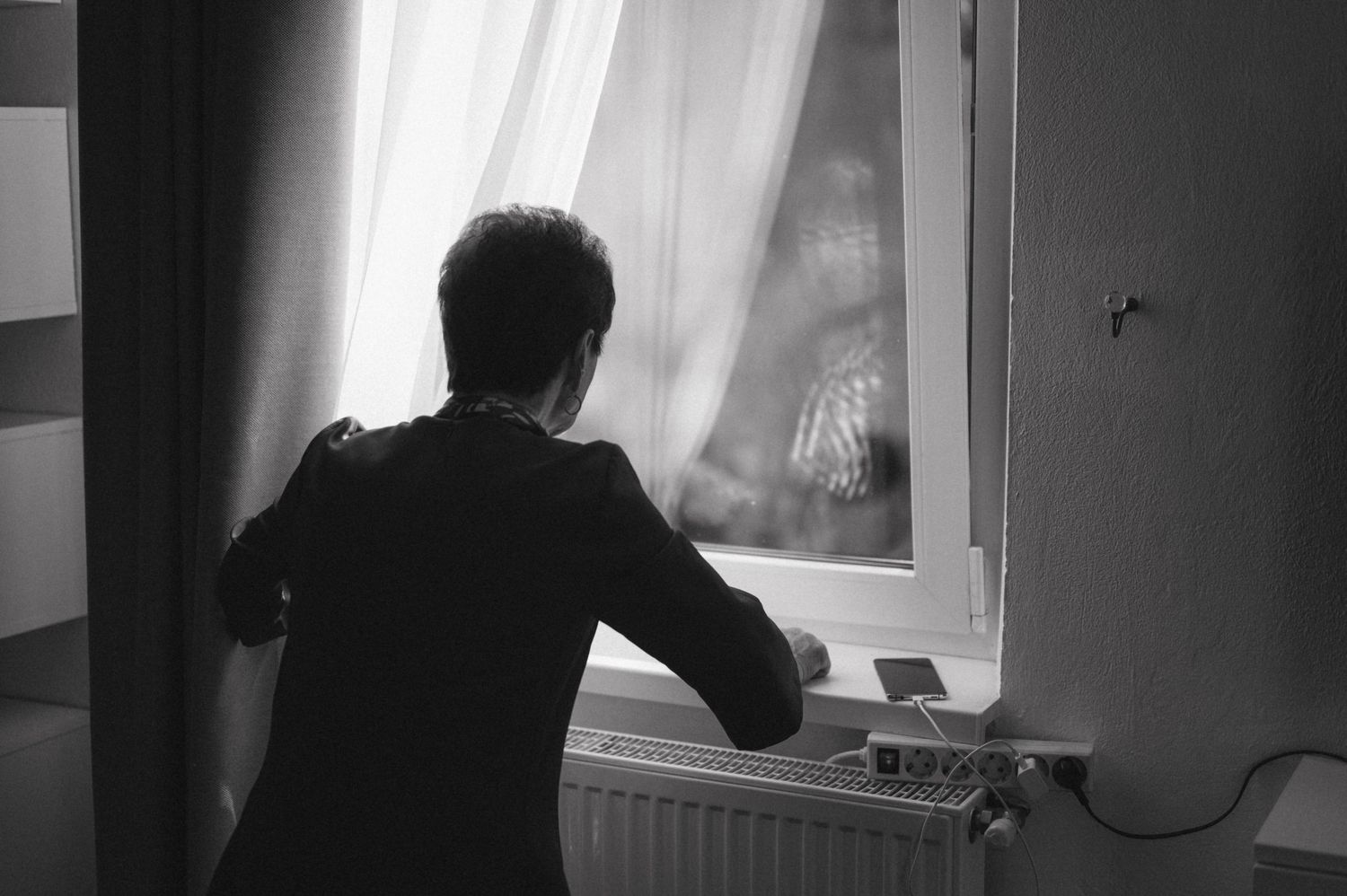 Silhouette of person looking through window with curtain and radiator.
