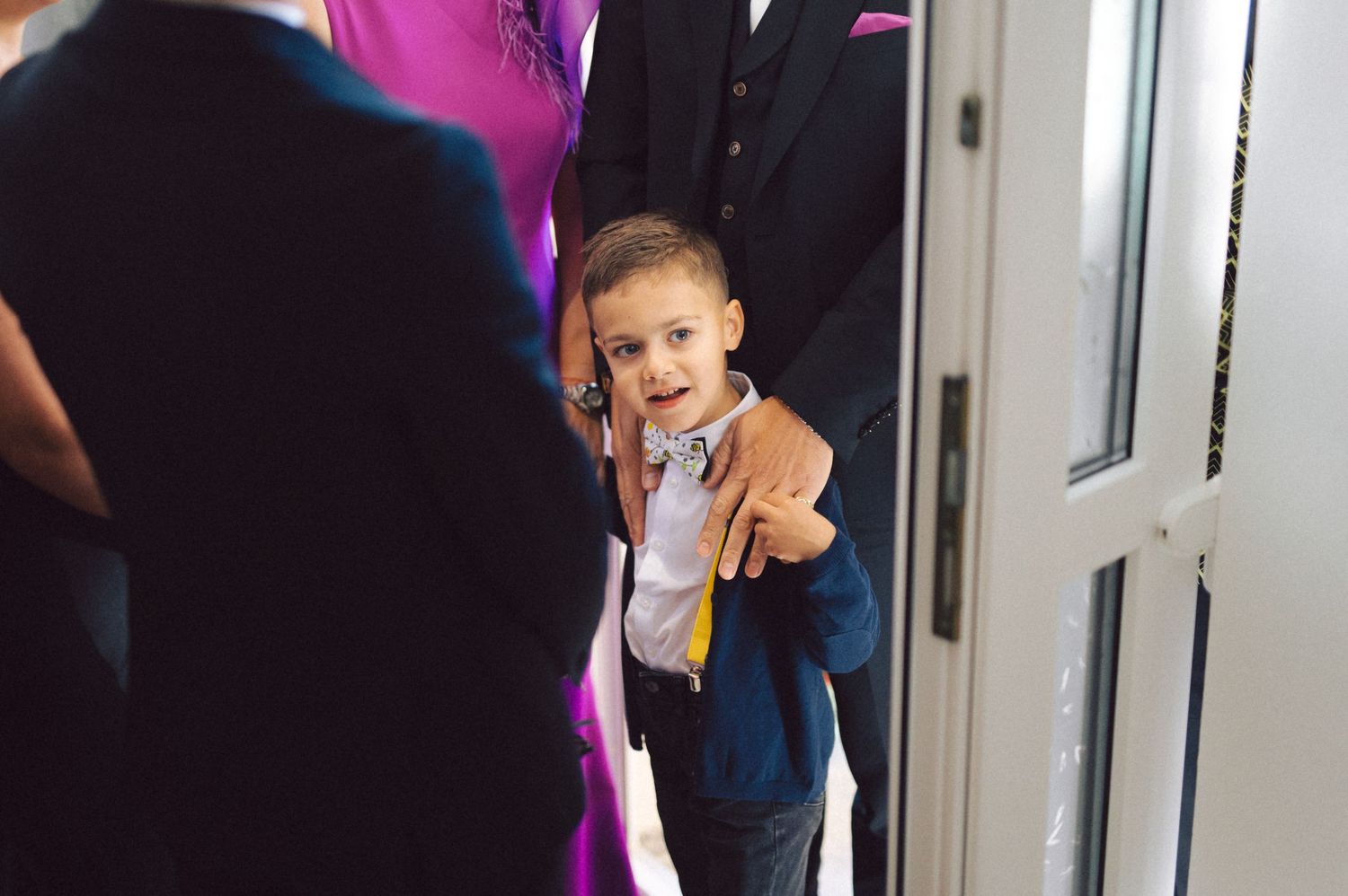 Young child in formal attire peeks through doorway during wedding celebration.