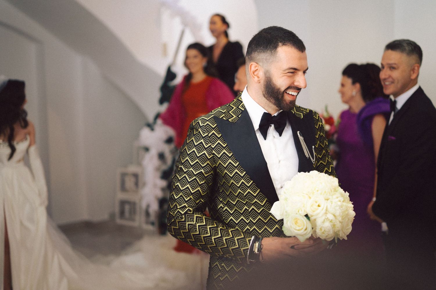 Guest in patterned tuxedo jacket carrying white flower bouquet at celebration.