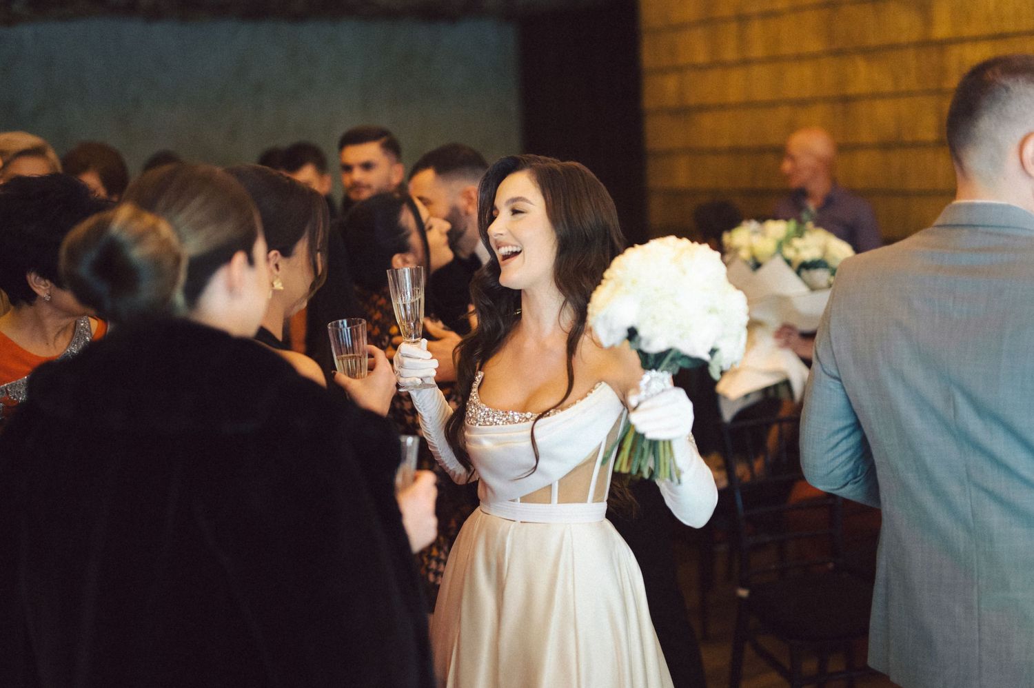 Joyful bride in white dress holds bouquet while celebrating with wedding guests at indoor reception.
