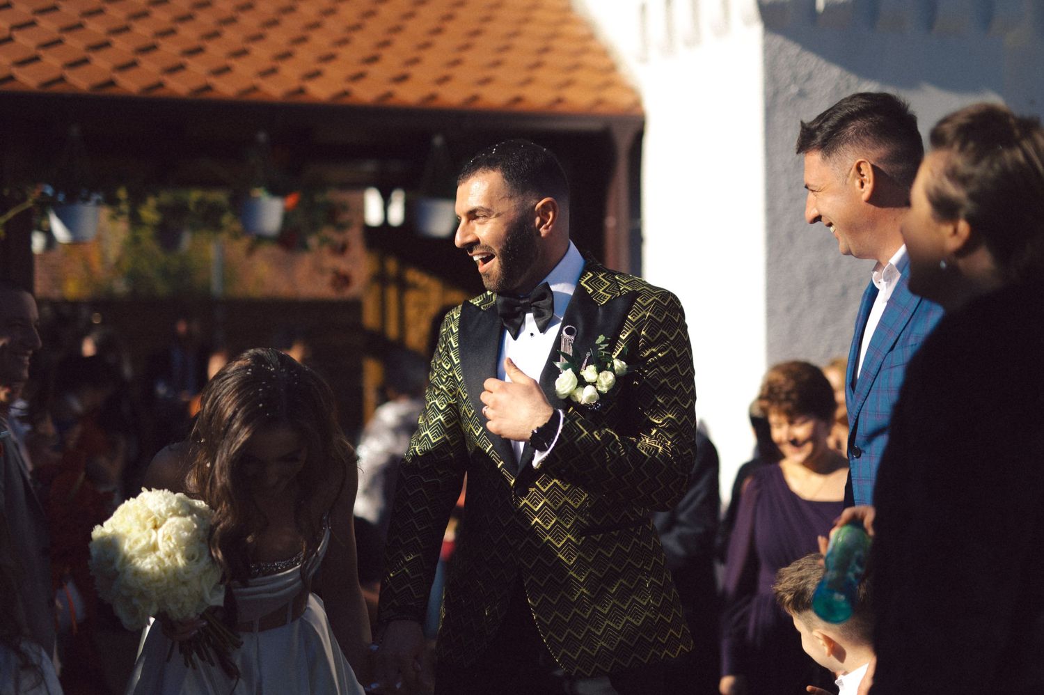 Wedding party gathers outdoors under a covered terrace during a sunny afternoon celebration.