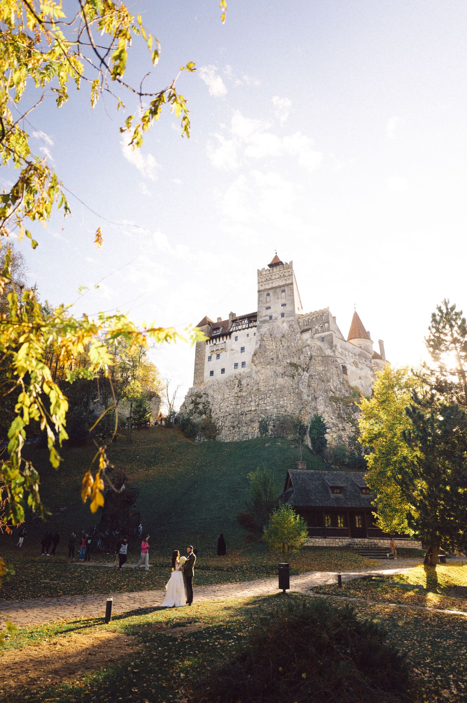 Bran Castle in Romania stands majestically on a hill during autumn with wedding couple in foreground.