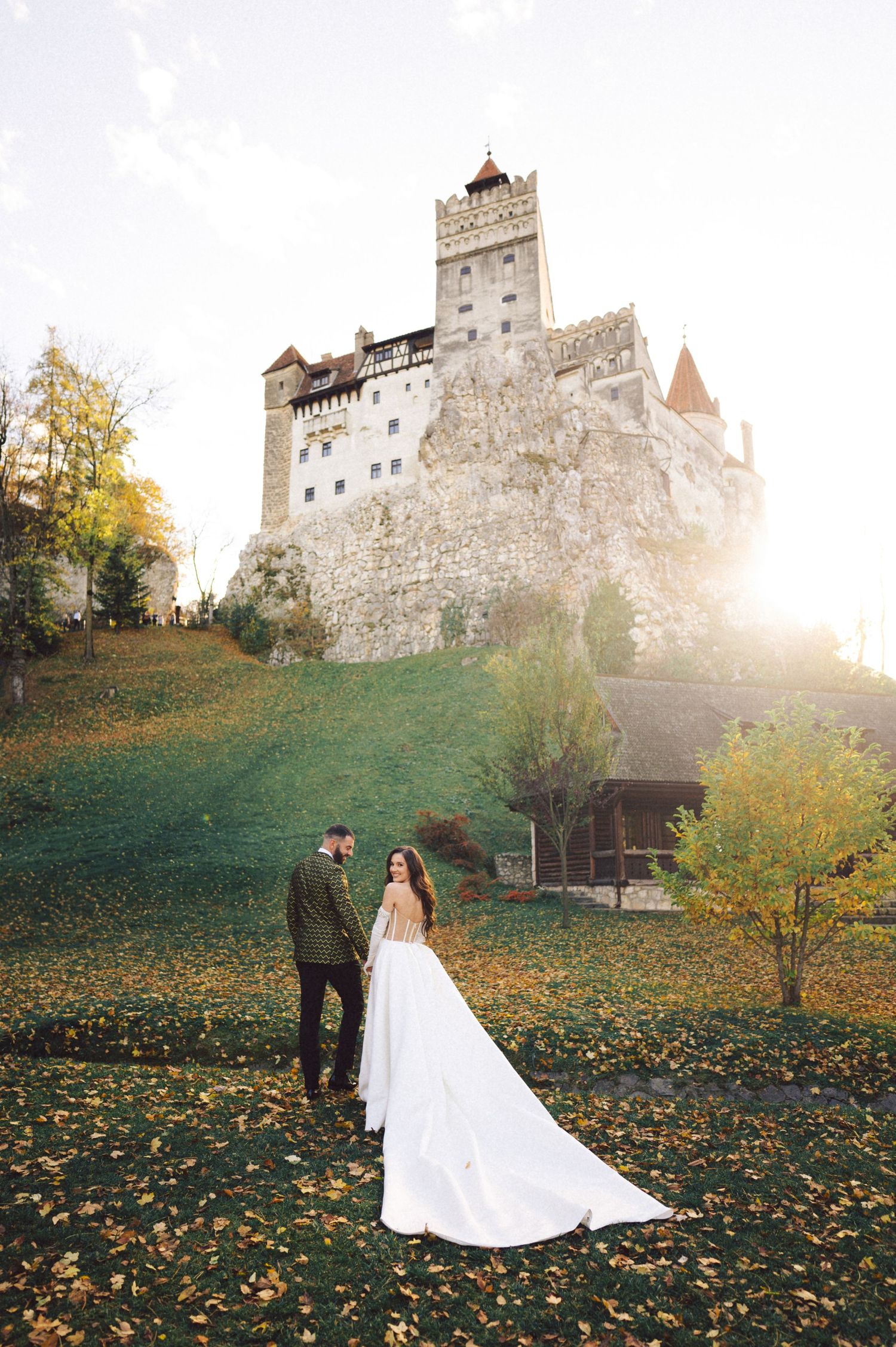 Newlyweds pose before historic Bran Castle with autumn foliage surrounding them.