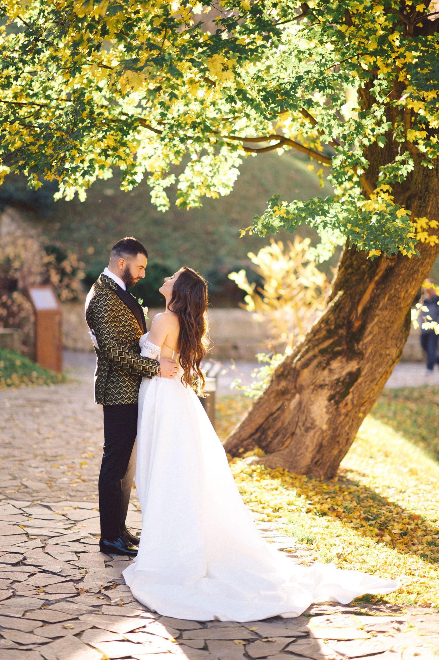 Wedding couple stands on stone path under golden autumn trees during sunset.