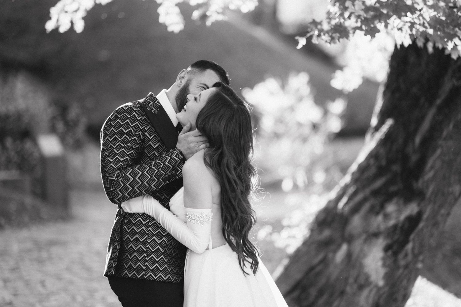 Romantic black and white wedding photo of a couple embracing under trees with soft bokeh lighting.