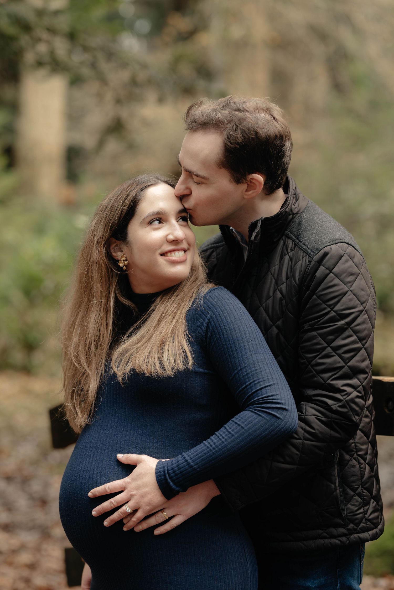 A couple shares an intimate moment outdoors while holding a pregnant belly wearing a blue dress and black quilted jacket.