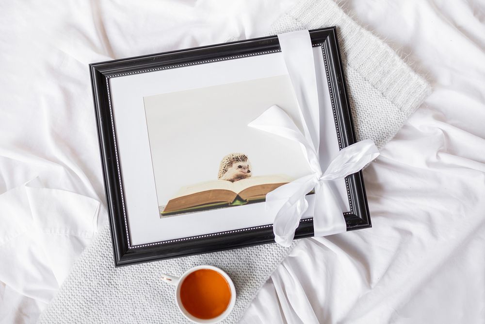 Flatlay of framed artwork with white ribbon, book, and tea cup on white textured fabric.