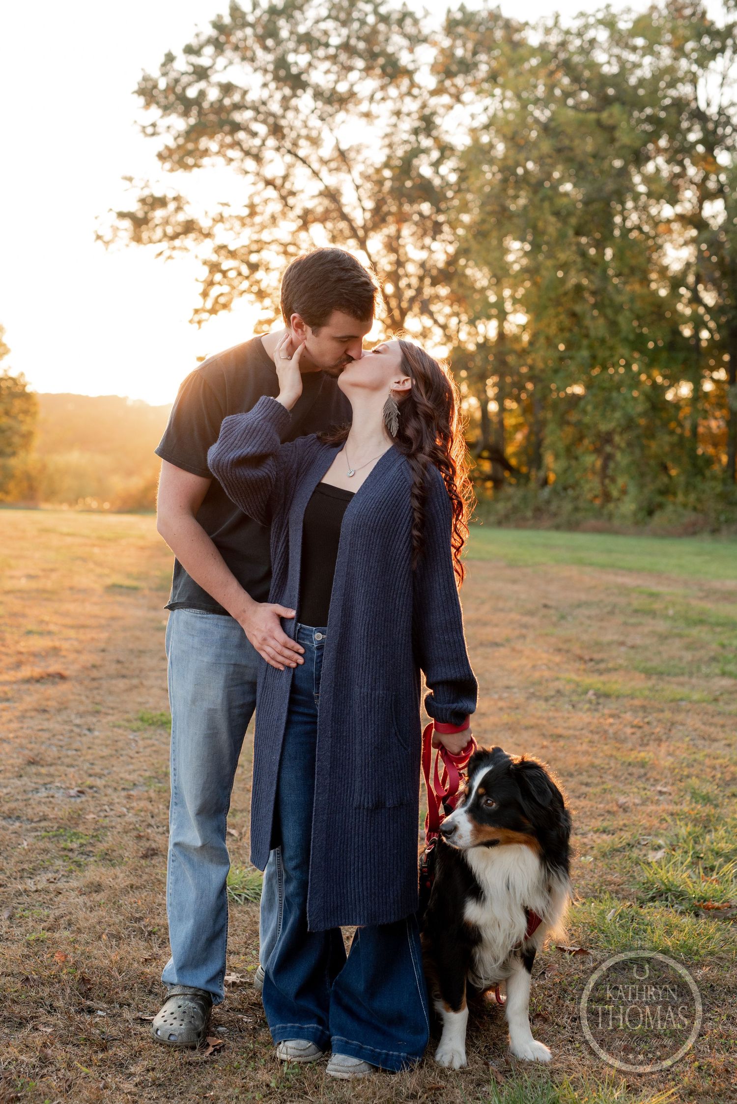 A couple shares a kiss while their Australian Shepherd companion stands beside them at sunset.