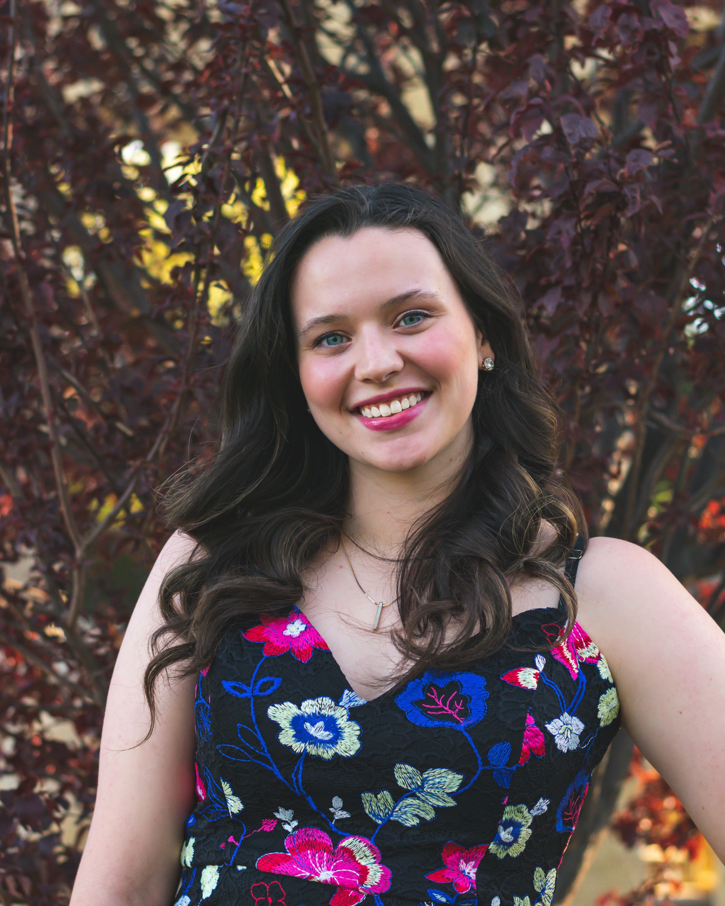 A bright smile stands out against dark burgundy foliage, wearing a vibrant floral dress with blue and pink blooms for graduate student
