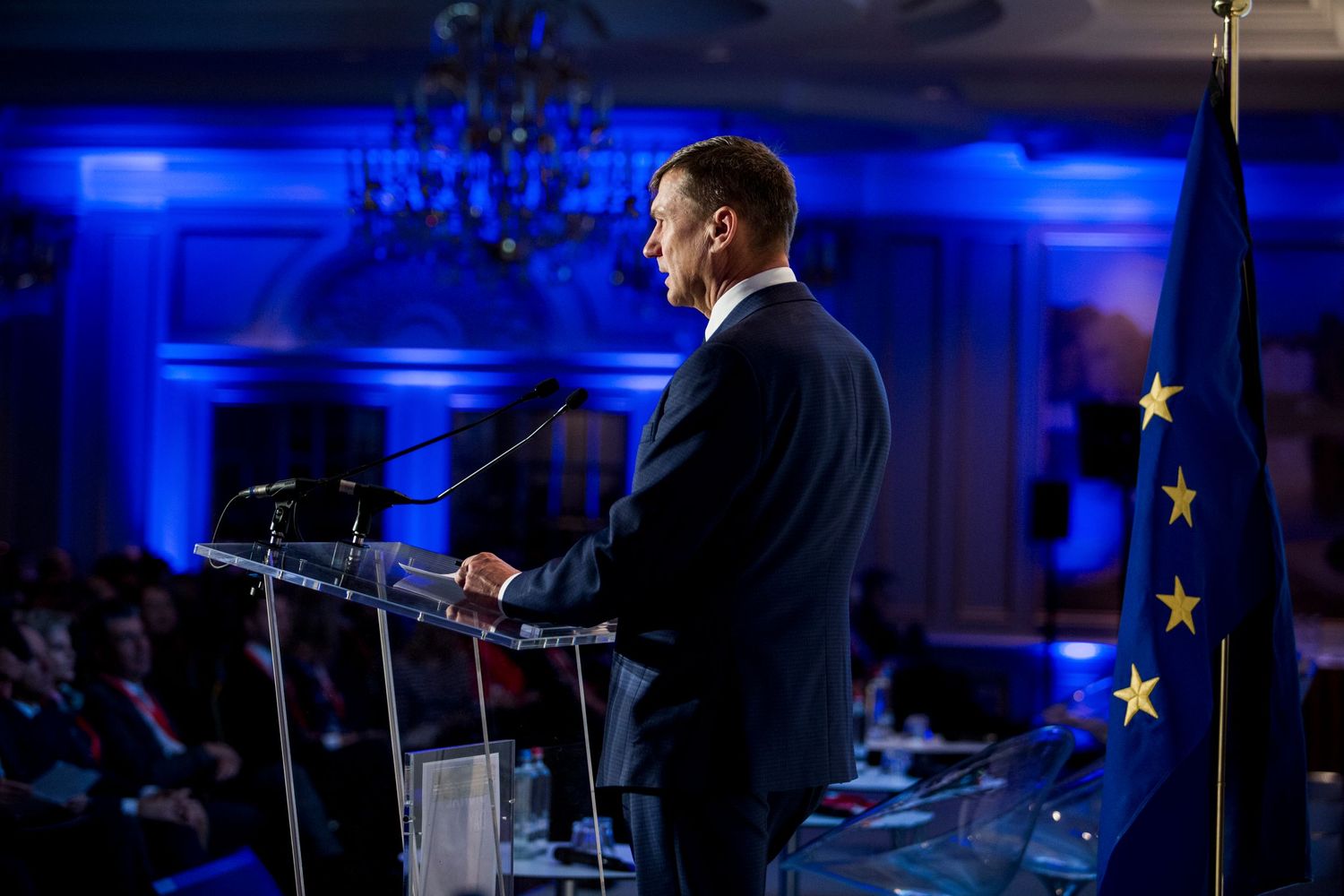 Speaker at podium with EU flag backdrop in dramatically lit blue room.