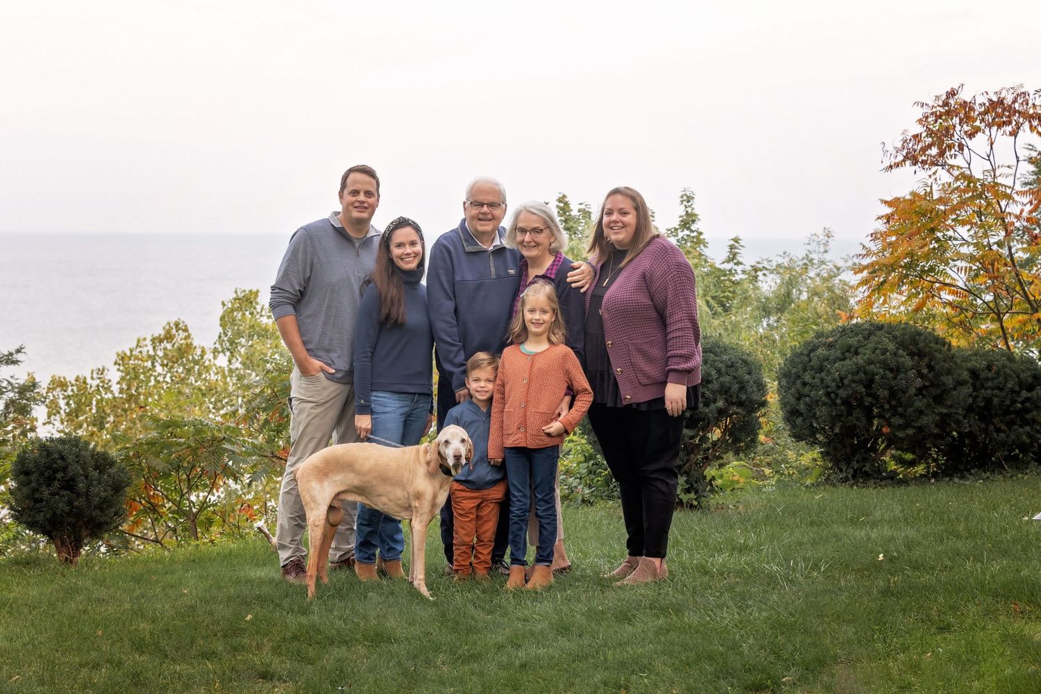 Multi-generational family and dog posing together on grassy hillside overlooking water.