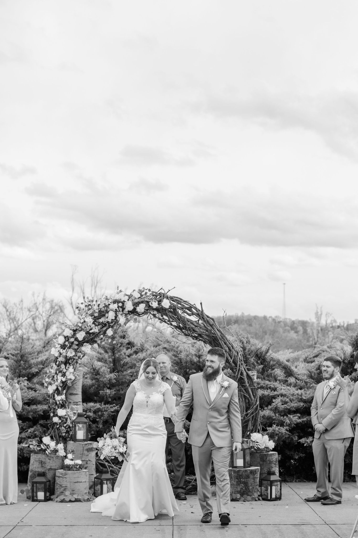 Black and white photo of a wedding ceremony under a decorated arch with guests seated outdoors.