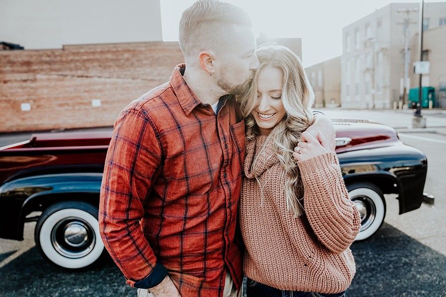 A romantic couple poses against a vintage car on an urban street.