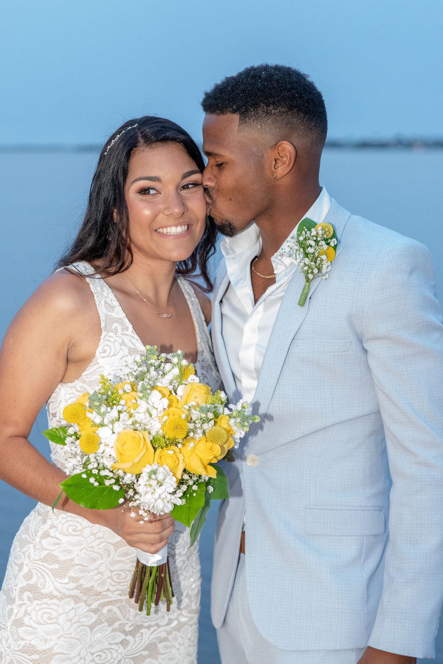 Romantic beach wedding portraits with yellow bouquet against light blue sky background.