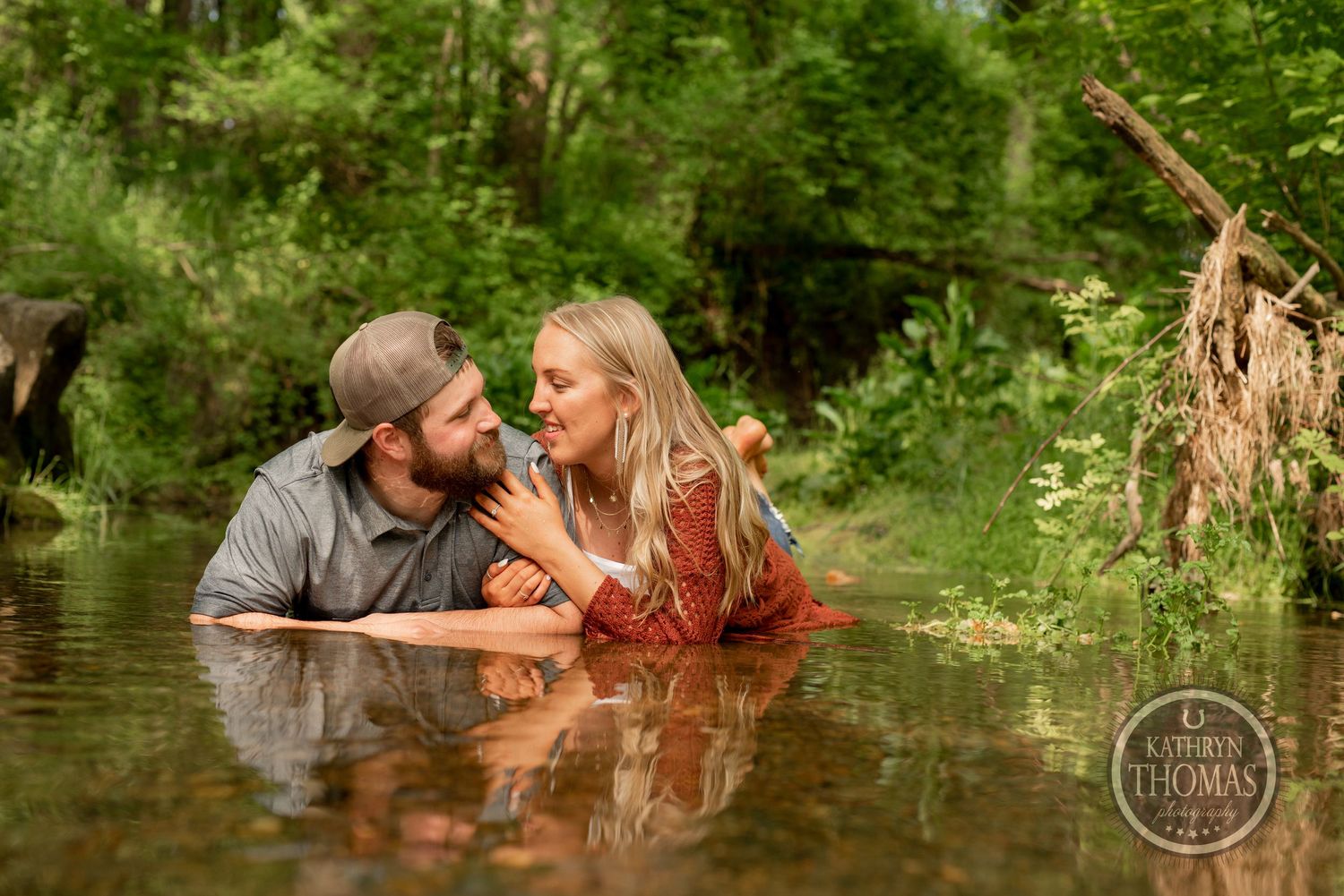 A romantic couple shares an intimate moment while partially submerged in a creek surrounded by lush greenery.