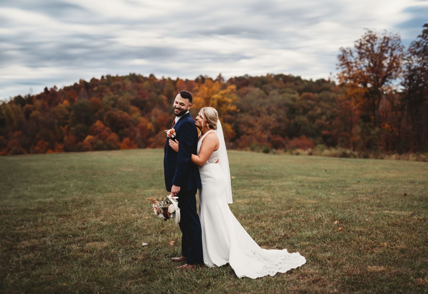 Bride and groom embrace during their autumn wedding in an open field surrounded by fall foliage on a cloudy day.
