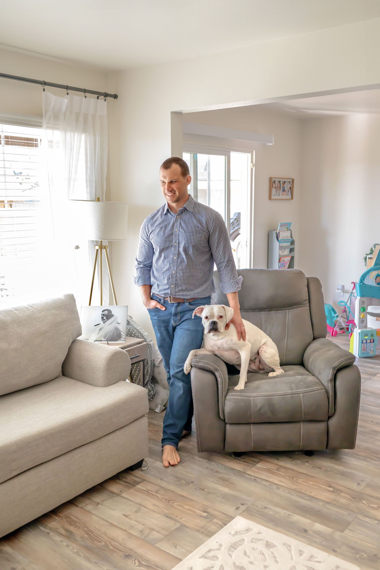 Dad stands next to the recliner with one hand gently petting the white boxer mix dog, who is sitting in the recliner. Include your pets!