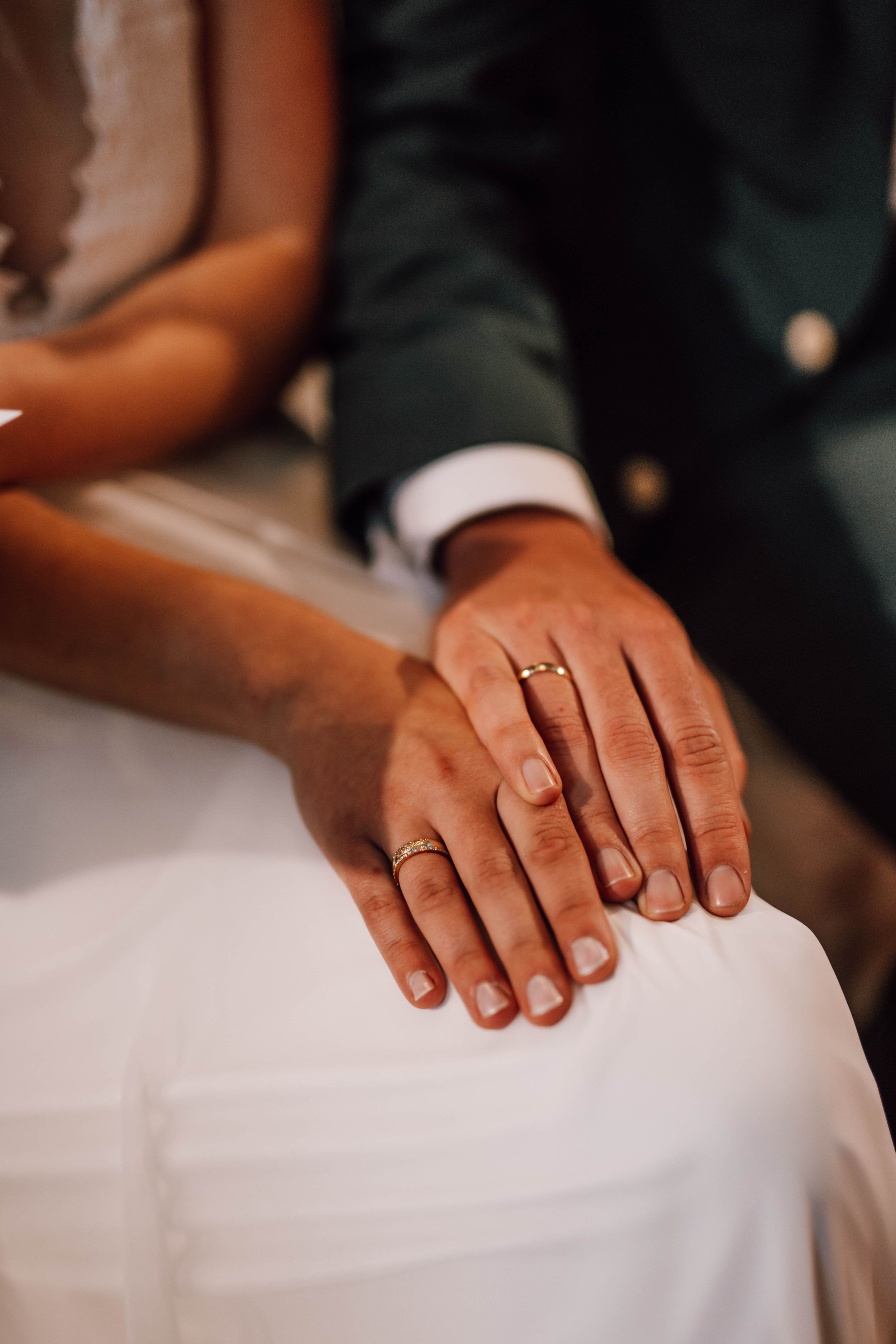 Close-up of newlyweds' hands with wedding rings resting on white dress fabric.