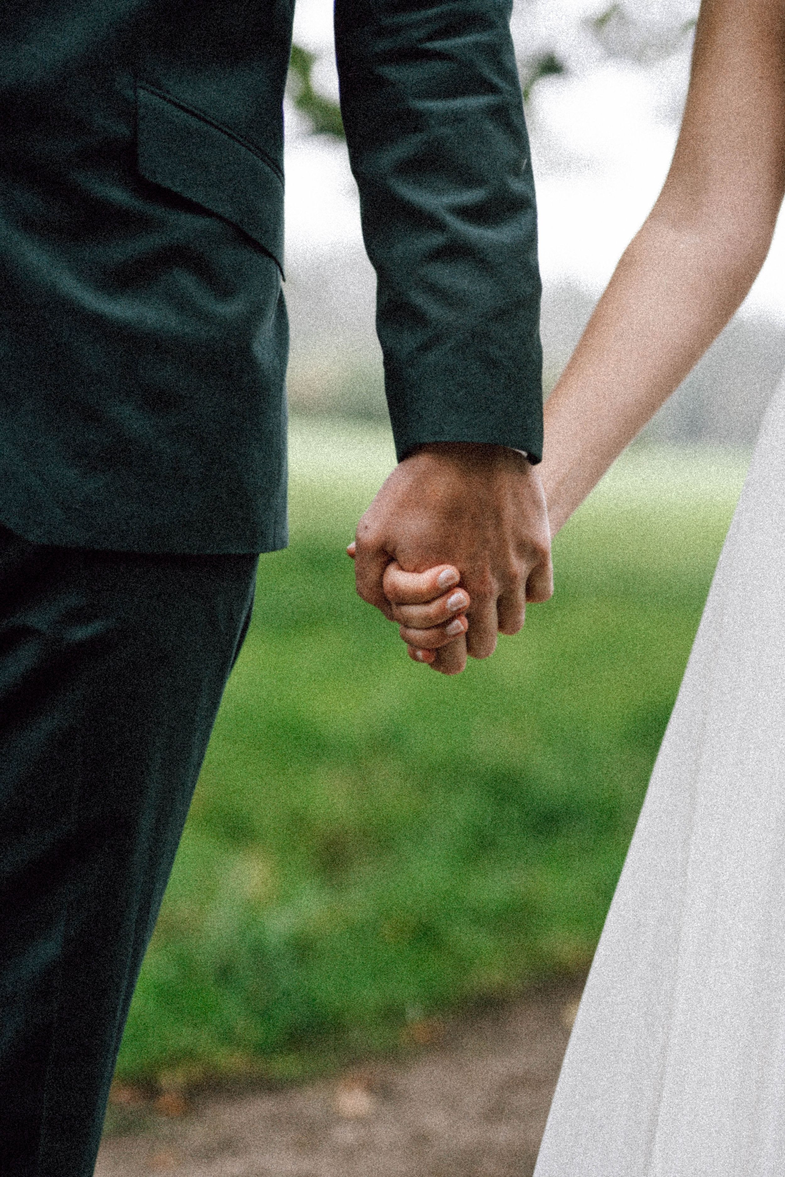Close-up of couple holding hands in formal wedding attire against green background.