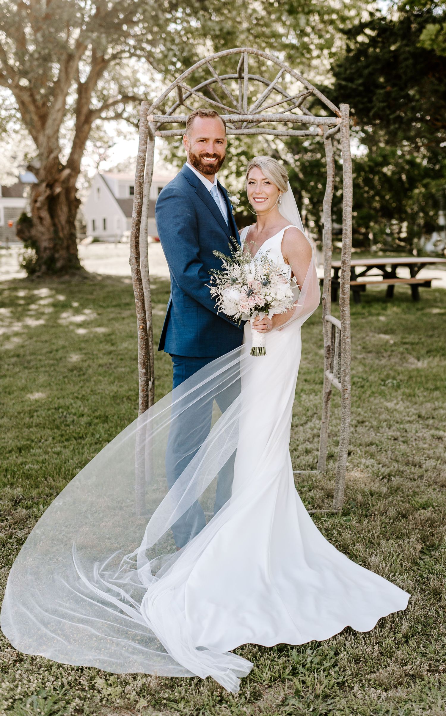 Couple poses under rustic wooden arch with flowing white veil in garden setting.