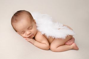 Sleeping newborn wearing white angel wings against neutral backdrop.