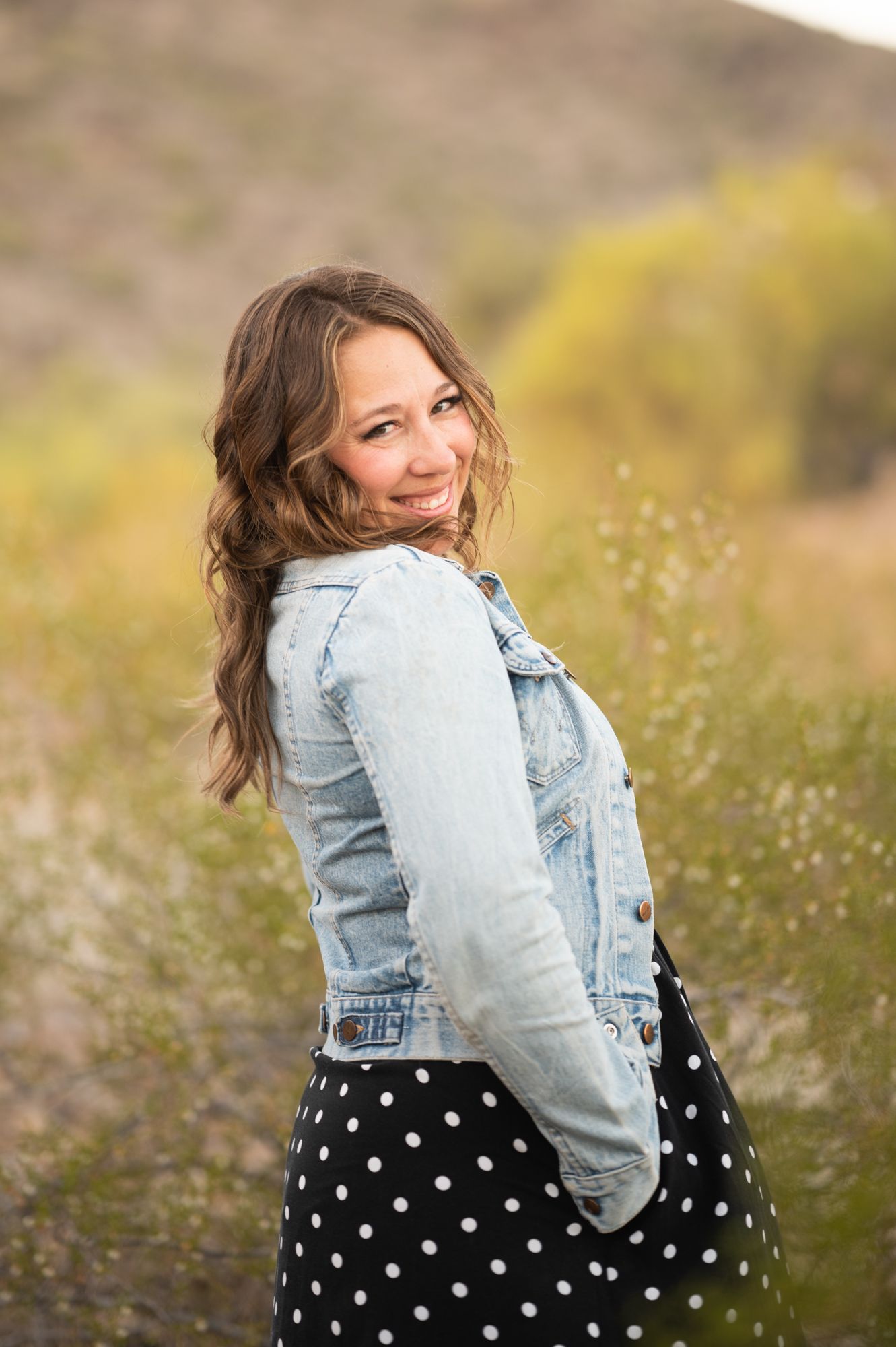woman with long wavy brown hair smiling over her shoulder in the desert with jean jacket