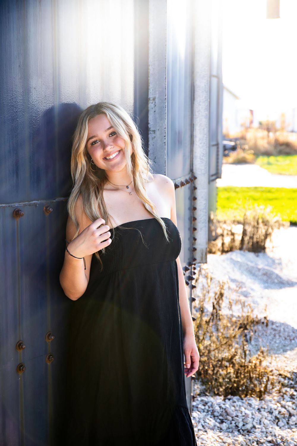 Sun-drenched fashion portrait featuring black dress and natural styling against metal wall.