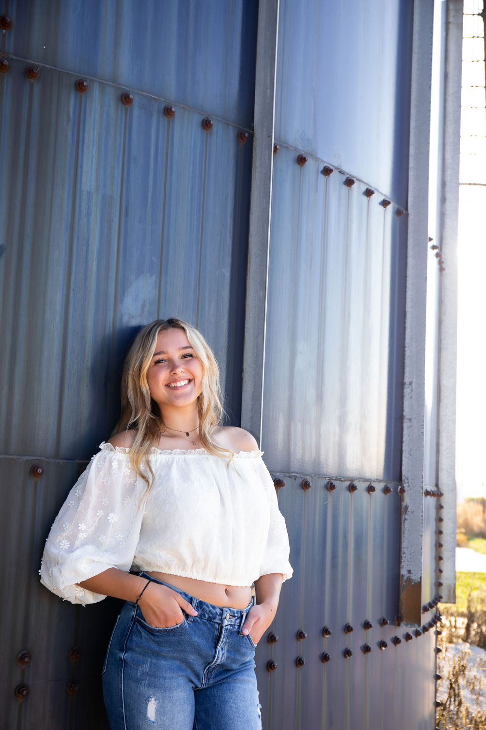 A stylish portrait against an industrial metal wall featuring a white off-shoulder top and distressed jeans.