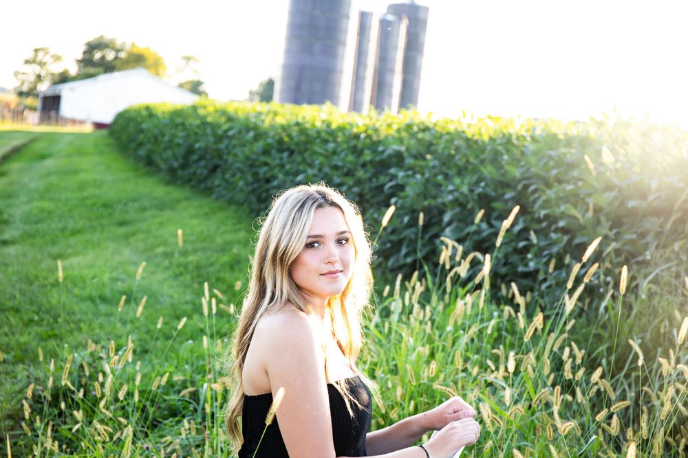 Outdoor portrait in a black dress sitting by tall silos and wildflowers during golden hour sunset.