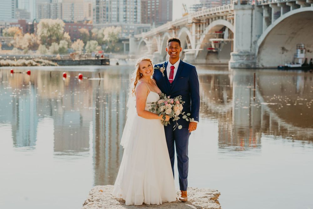 Bride and groom standing on rocks by a river with a city bridge backdrop.