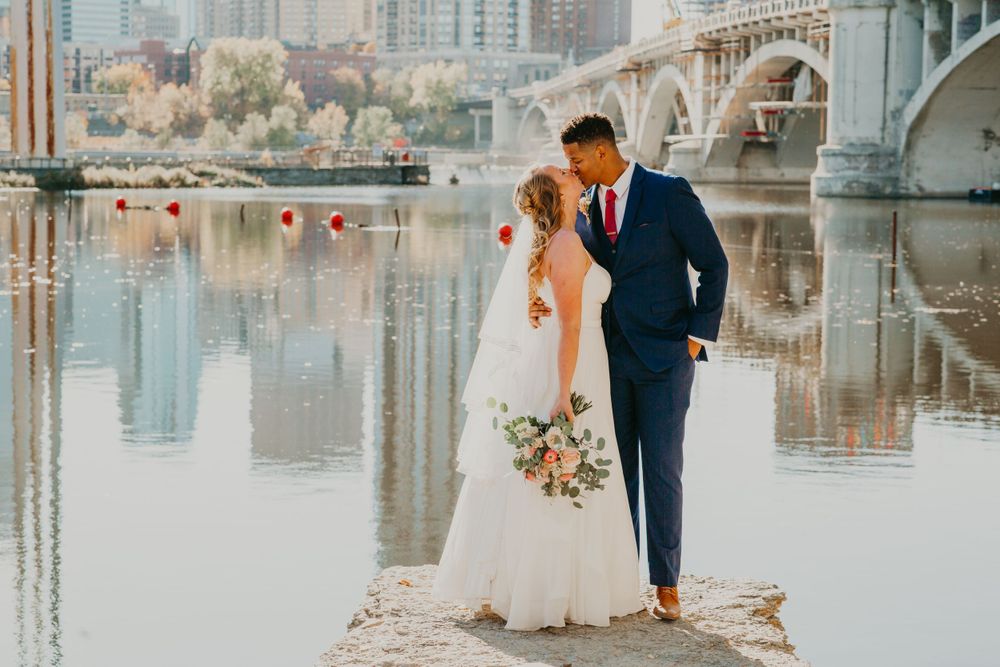 Bride and groom sharing a kiss on rocks by a river with a bridge backdrop.
