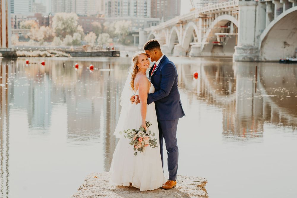 Bride and groom share a romantic moment on a rocky shore with a bridge and river in the background.