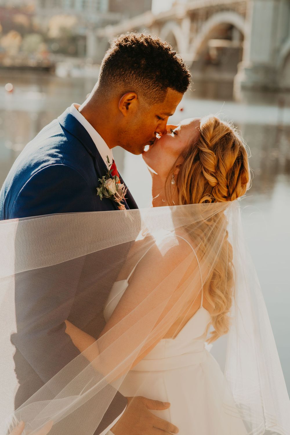 Bride and groom share a kiss under a flowing veil with a sunlit river and bridge backdrop.