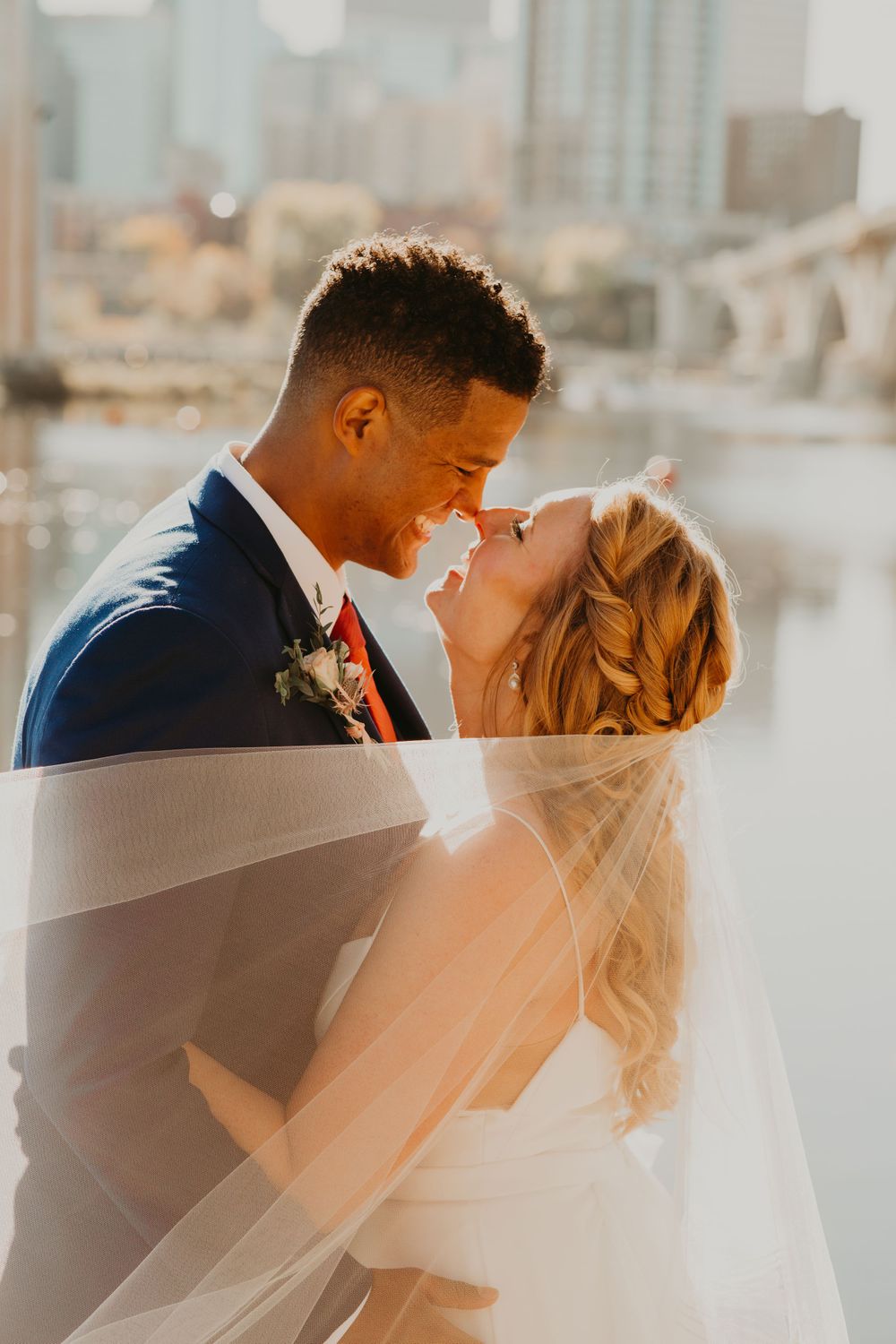 Smiling bride and groom nearly kiss under a sheer veil with a city skyline and river behind them.
