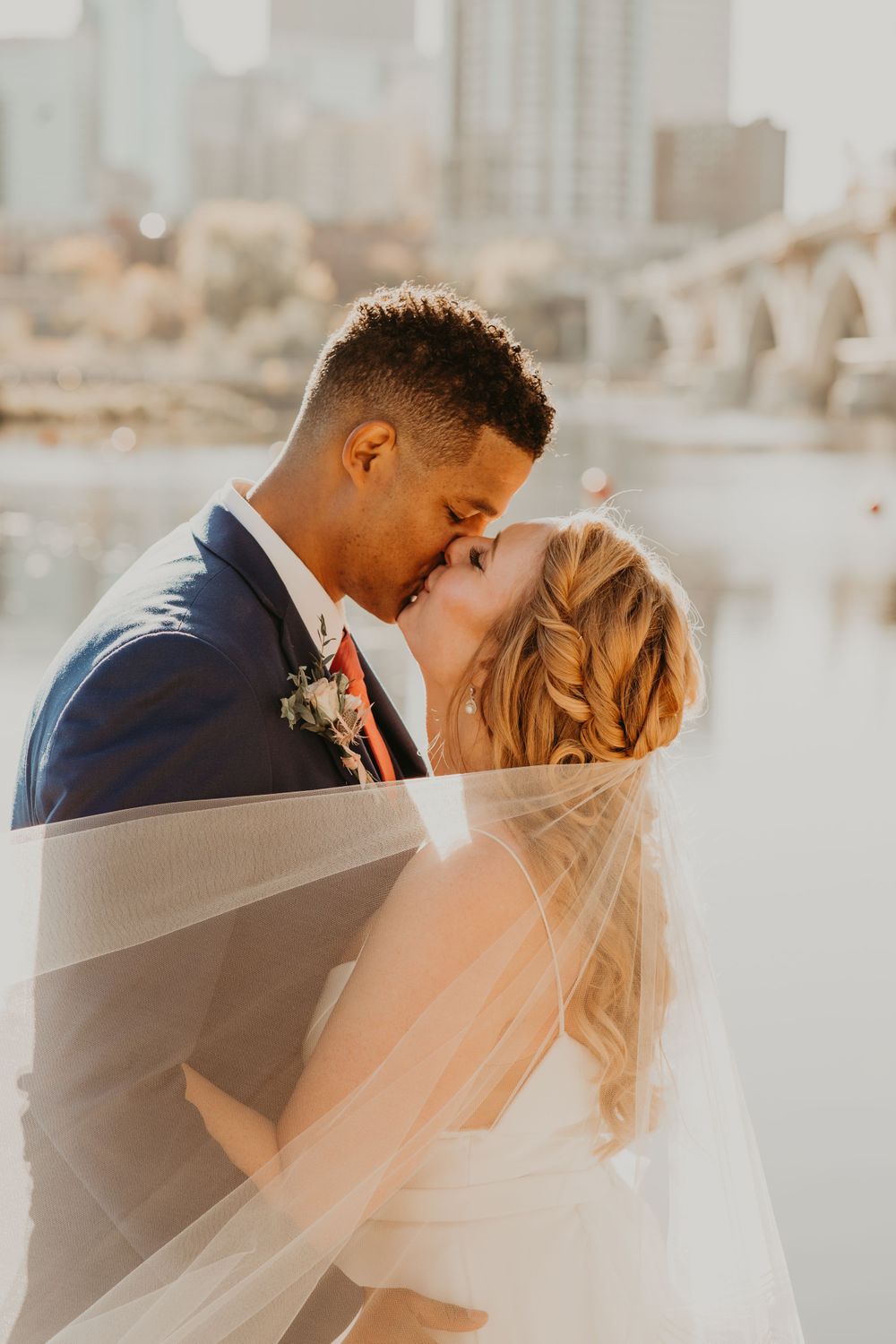 Bride and groom share a passionate kiss under a veil with a city skyline and river in the background.