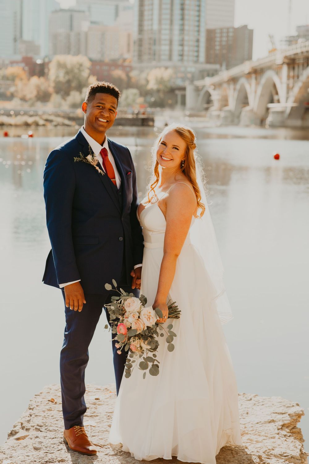 Bride and groom pose together by a river holding a floral bouquet with a city bridge in the background.