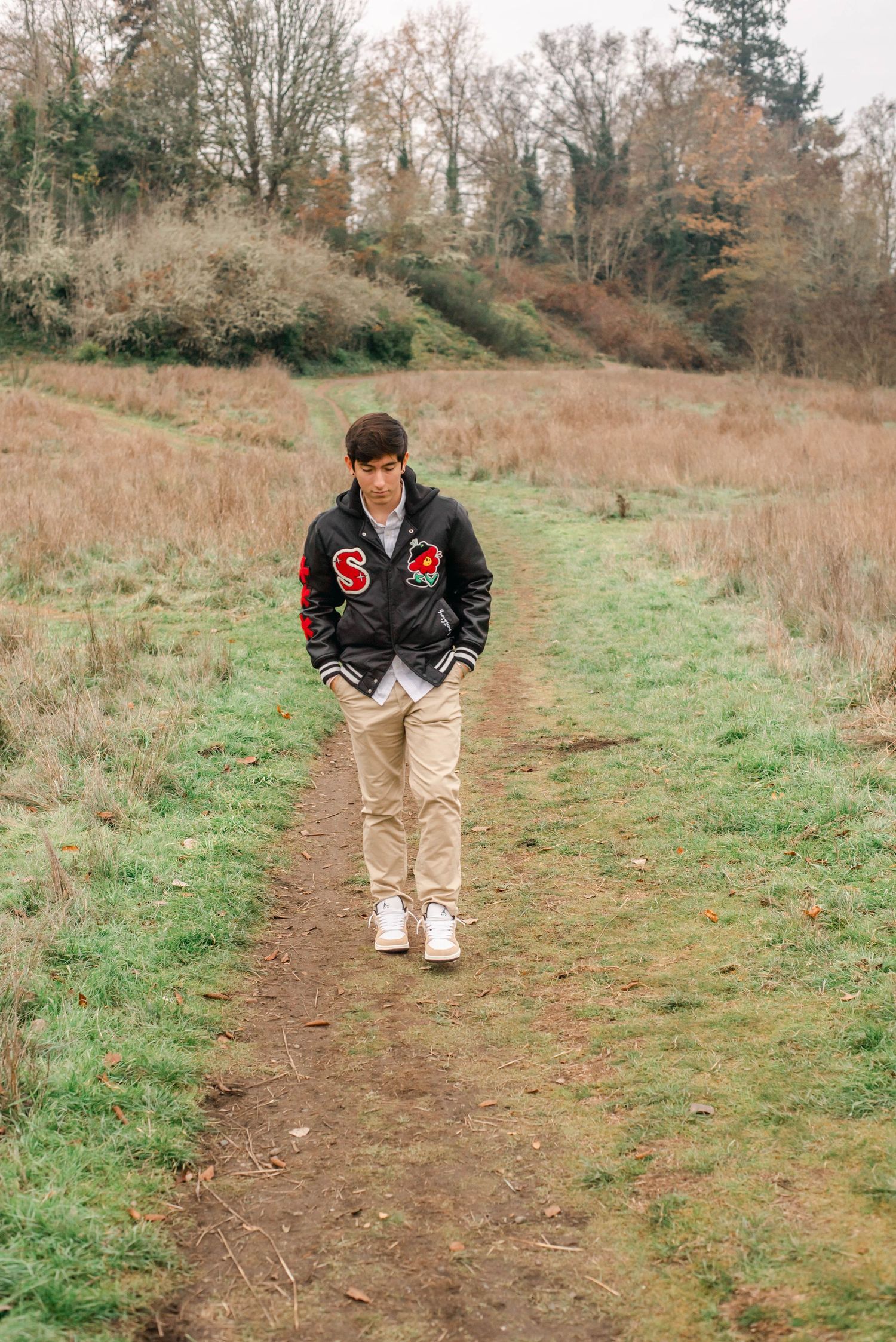 Person wearing a black jacket and khaki pants poses on a dirt path surrounded by autumn foliage.
