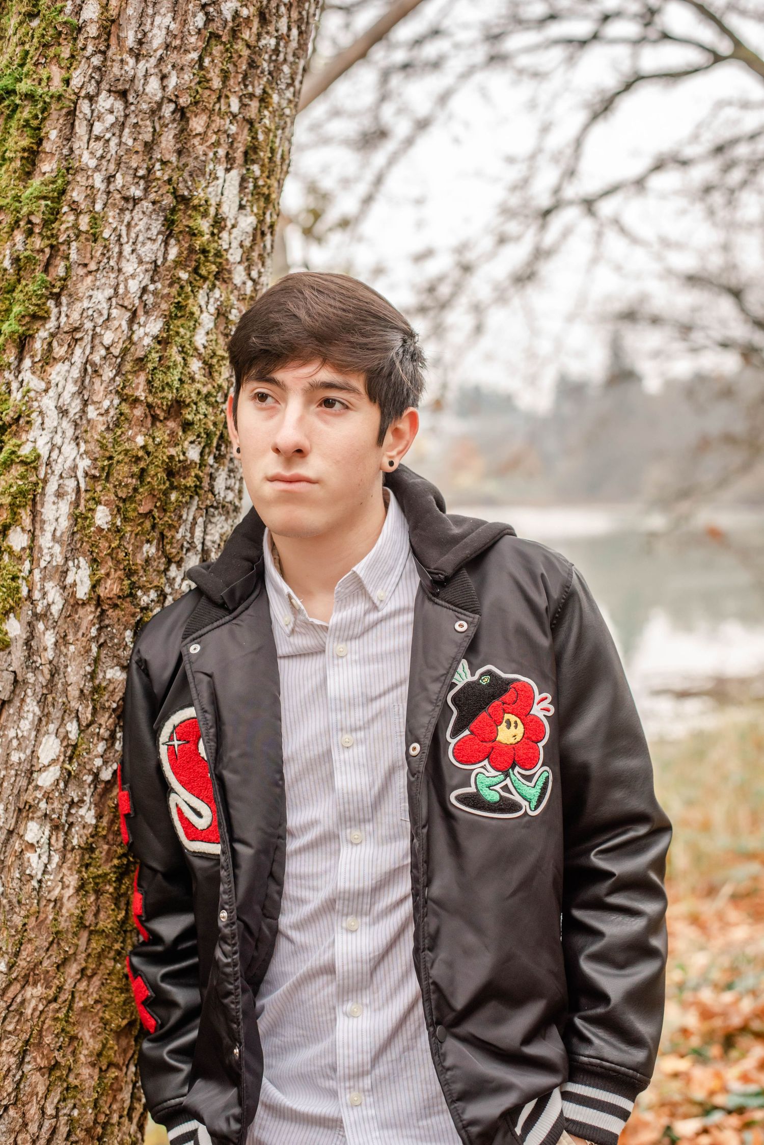 Portrait series near tree trunk wearing black jacket with decorative patches.