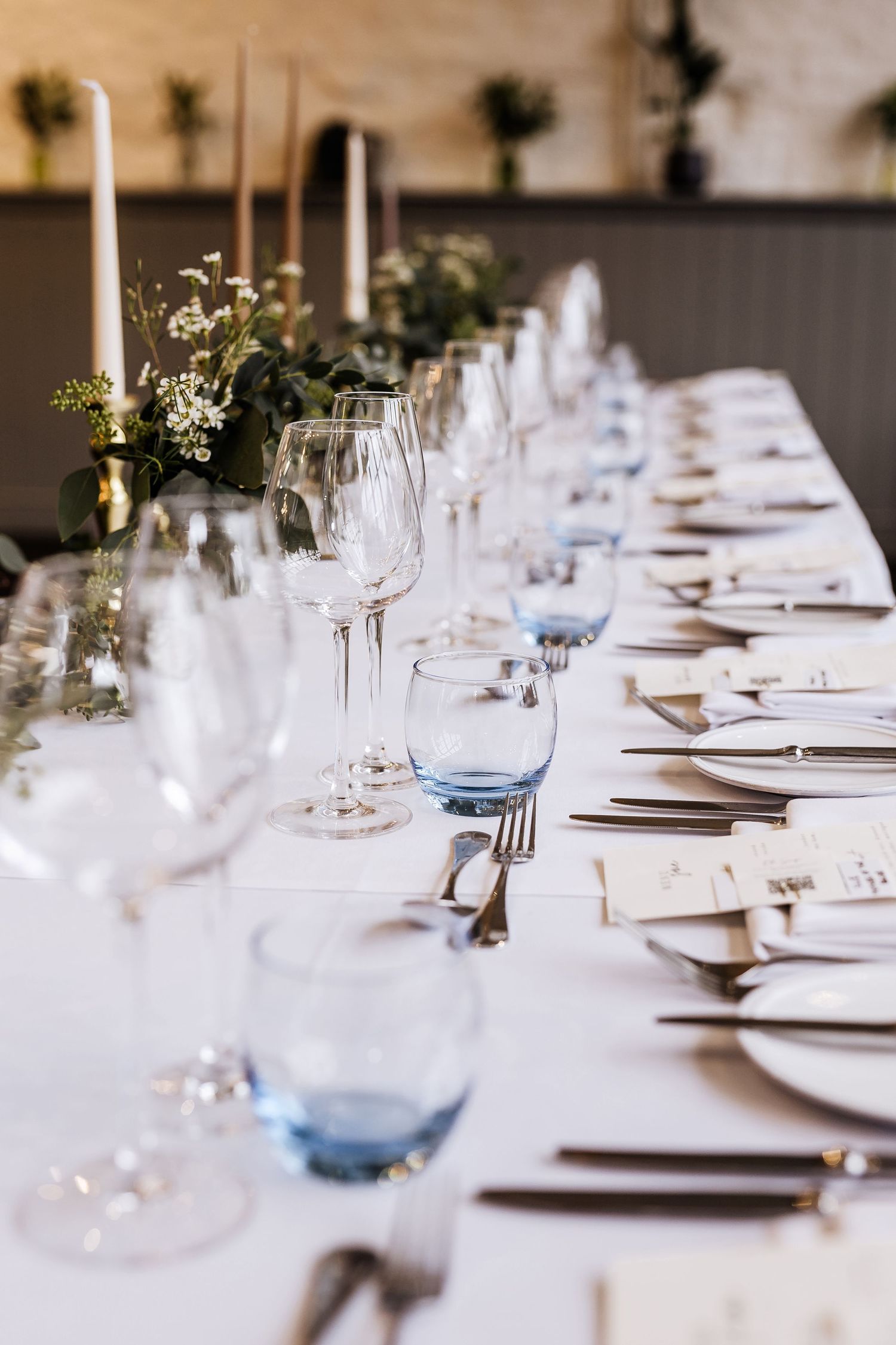 A formal table setting with blue glassware, white linens, and simple greenery centerpieces.