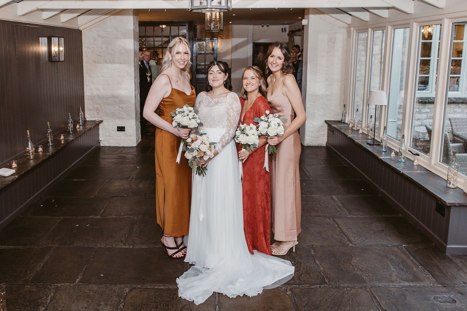 Wedding party poses in elegant dresses including rust, red and blush tones against stone wall interior.