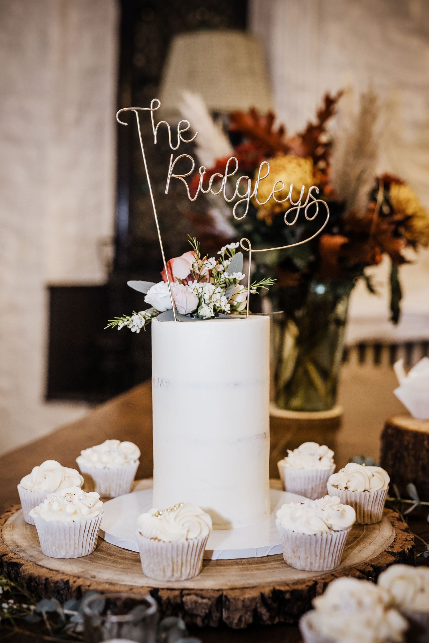A minimalist white wedding cake display with cupcakes on a rustic wood slice with wire text topper.