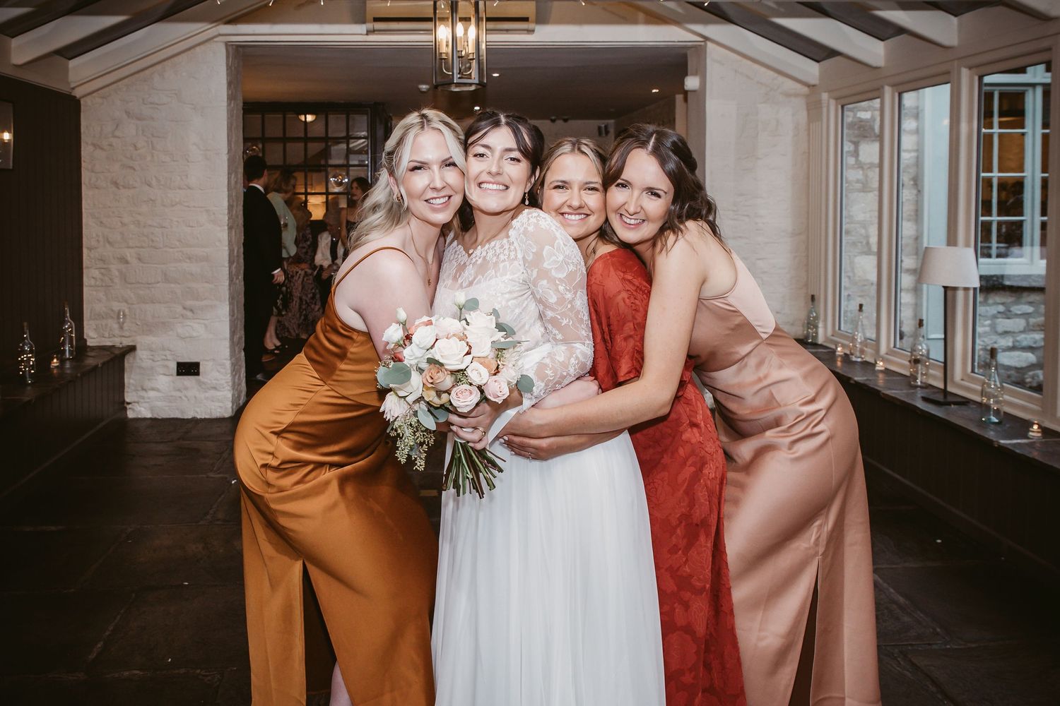 Bridal party wearing silk dresses in autumn colors embrace in joyful group pose.